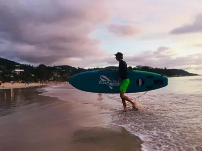 a man walking on the beach with a surfboard at Recommend a warm Thai style private swimming pool villa with convenient traffic and living, walking 700 meters to the the beach in Kamala Beach
