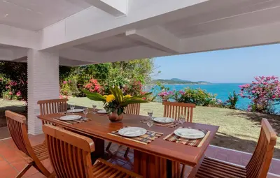a wooden table with chairs on a patio with the ocean at Baan Khunying - Secluded Phuket Beachfront Villa in Rawai Beach