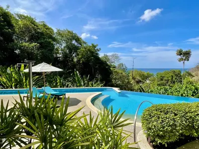 a blue swimming pool with an umbrella and the ocean at Kata Seaview Serenity - Luxury 1BR Apartment in Karon Beach