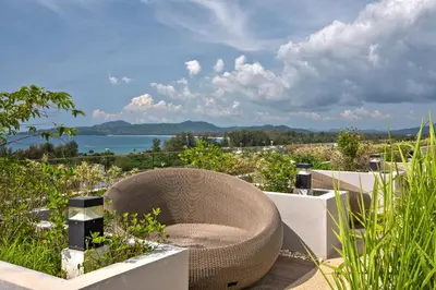 a rattan chair sits on a patio overlooking the water at Ocean Breeze 1-BR apartment, Sea View, Surin beach in Ban Lum Fuang