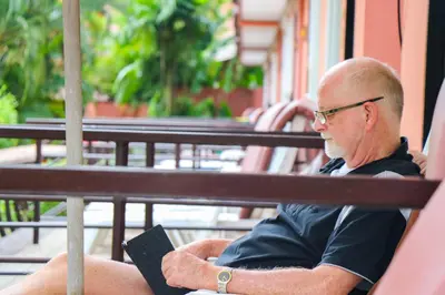 an older man sitting on a bench with a book at Andaman Seaside Resort Bangtao Beach in Bang Tao Beach