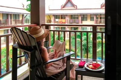 a woman sitting in a chair reading a book on a balcony at Diamond Cottage Resort & Spa in Karon Beach